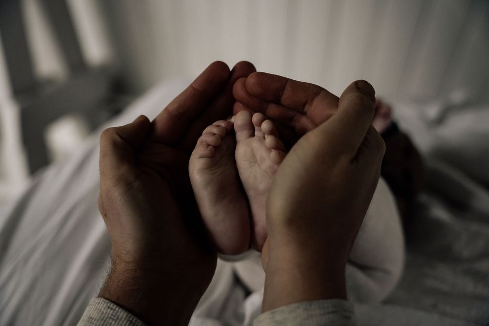 Adult hands gently holding tiny baby feet against white bedding in soft natural light.
