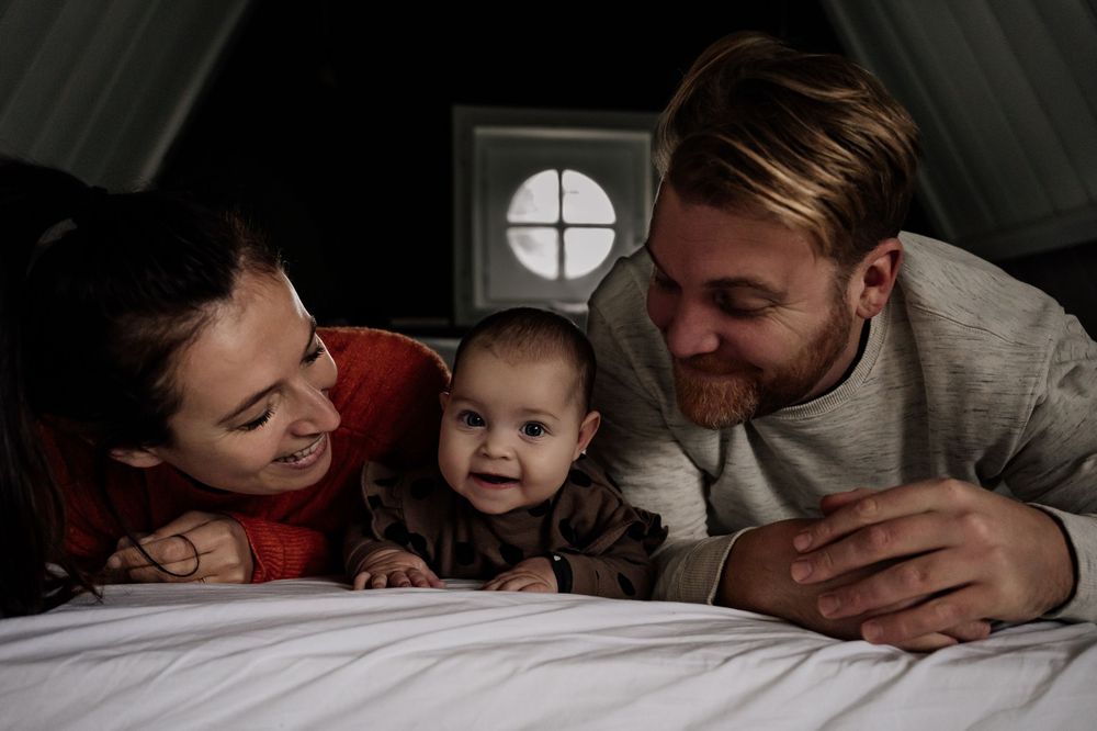A family lying together on white bedding sharing an intimate moment in dim lighting.