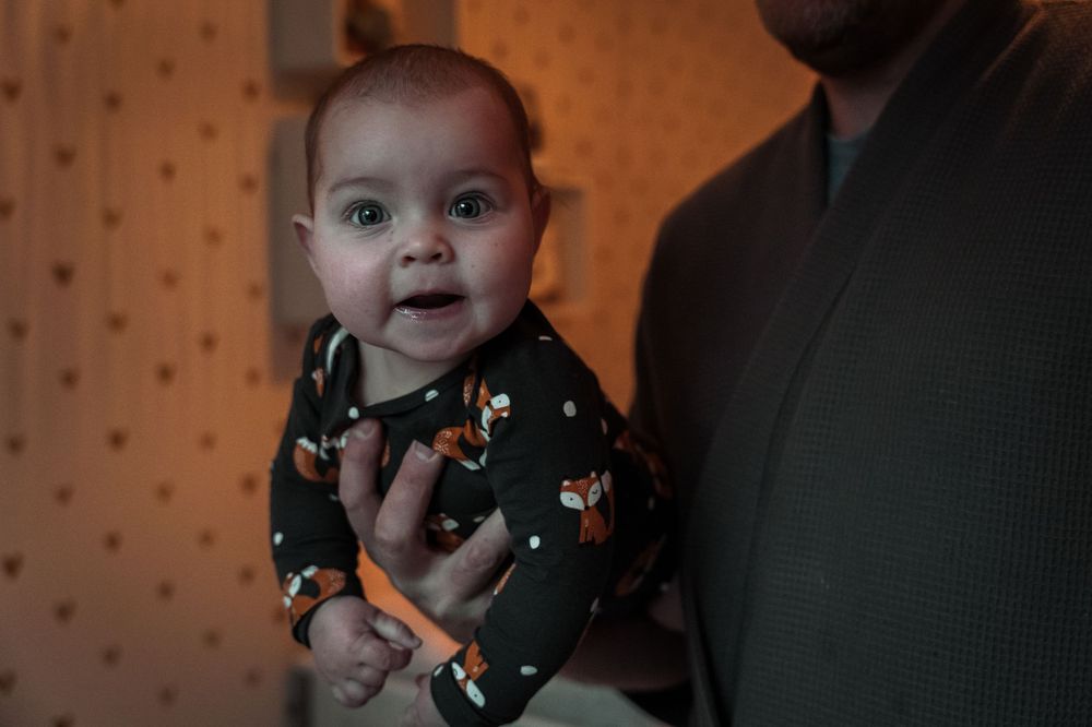 A baby wearing a dark green holiday outfit smiles while sitting in a cozy room with polka dot wallpaper.
