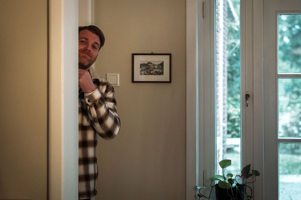 Someone wearing a plaid shirt peeks playfully from behind a doorway in a sunlit interior space.