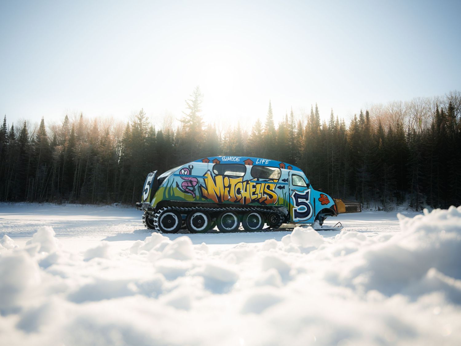 Colorful graffiti-wrapped snowcat vehicle parked on a snowy landscape with sunlit pine forest in the background.