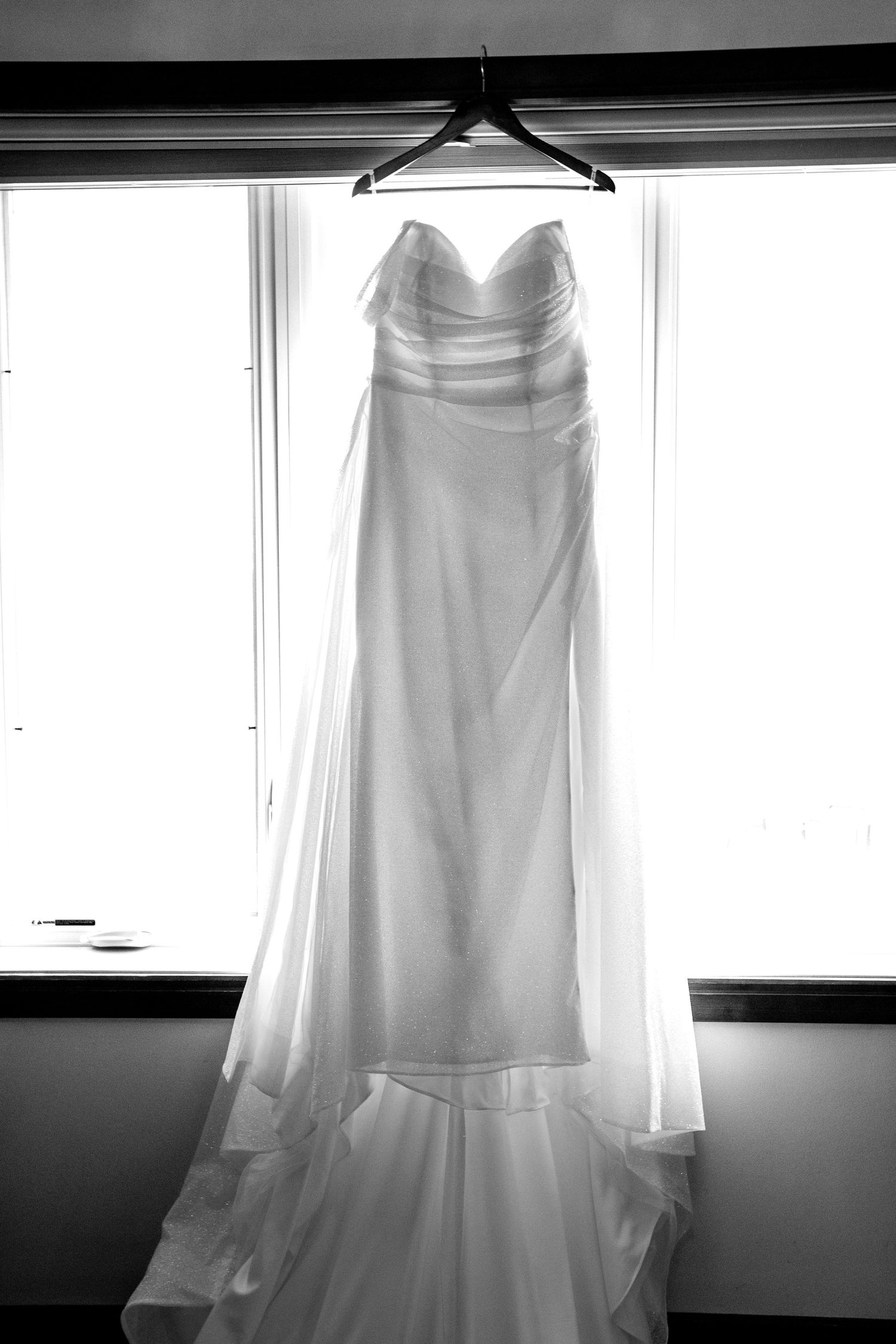 An elegant white wedding dress hangs from a black hanger against a window in dramatic black and white lighting.
