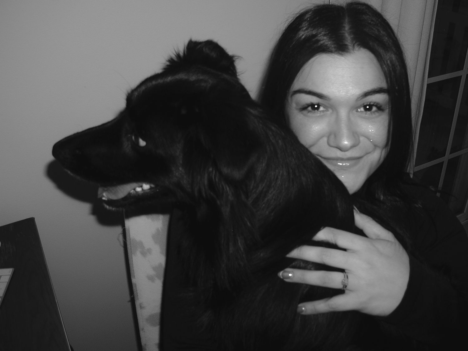 A black and white photo shows someone posing with a friendly black dog in a close embrace.
