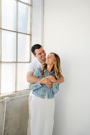 A couple shares an intimate embrace by a bright window, with her wearing a denim jacket and white dress.