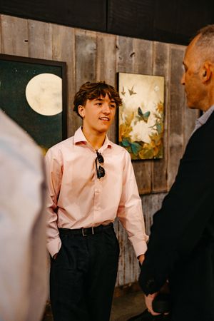 A child in a pink dress shirt and dark vest looks up while standing against a wall with artwork.