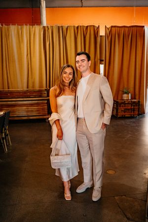 A couple poses together at an indoor venue, wearing elegant beige and white formal attire against brown curtains.