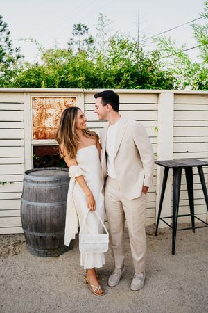 Romantic couple in white and beige outfits posing against white fence with rustic wine barrels in outdoor setting.