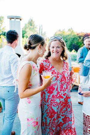Wedding guests in red and pink floral dresses socialize and enjoy cocktails at an outdoor summer reception.