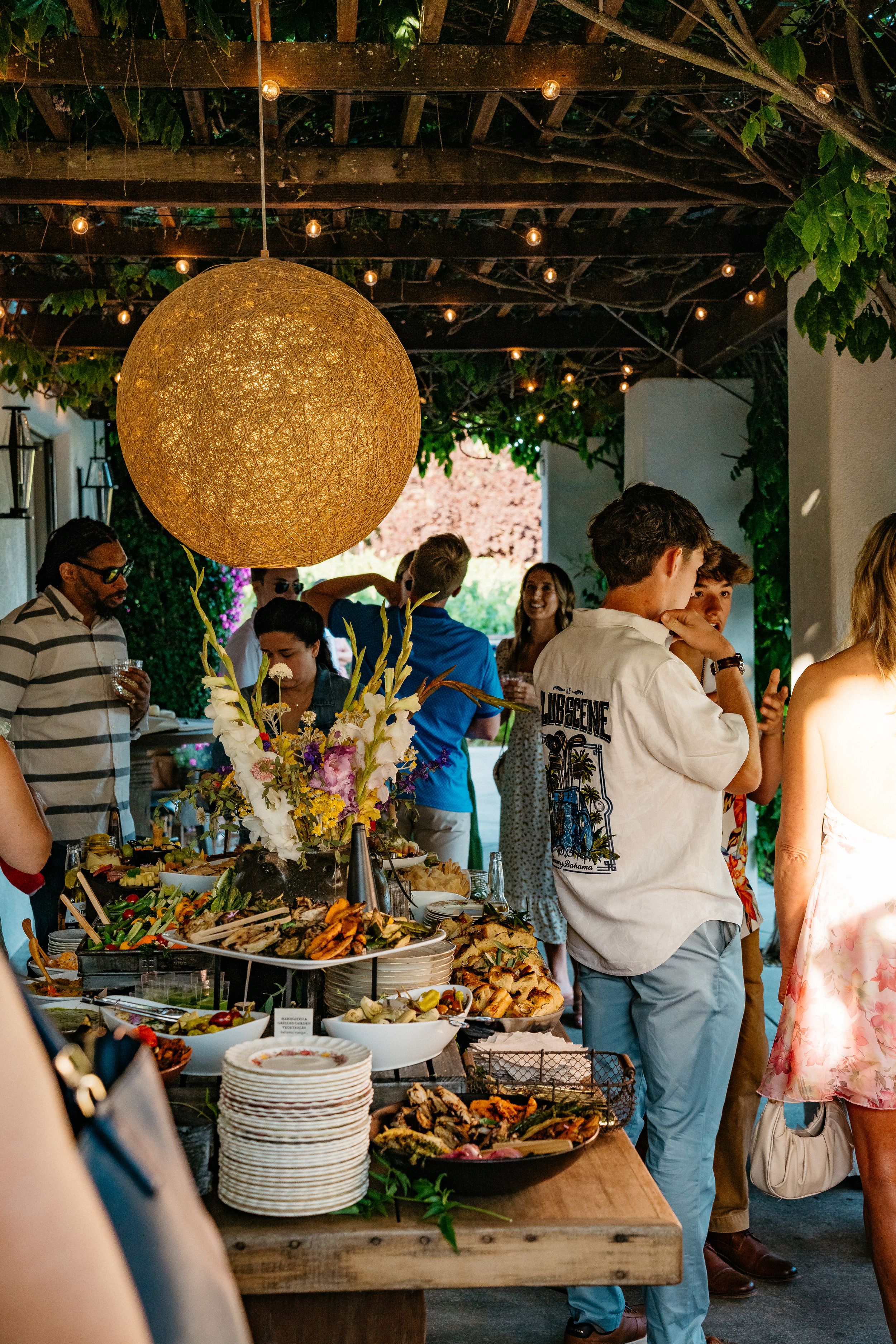 People gather at a rustic buffet table under warm lighting with decorative hanging lamps and greenery overhead.