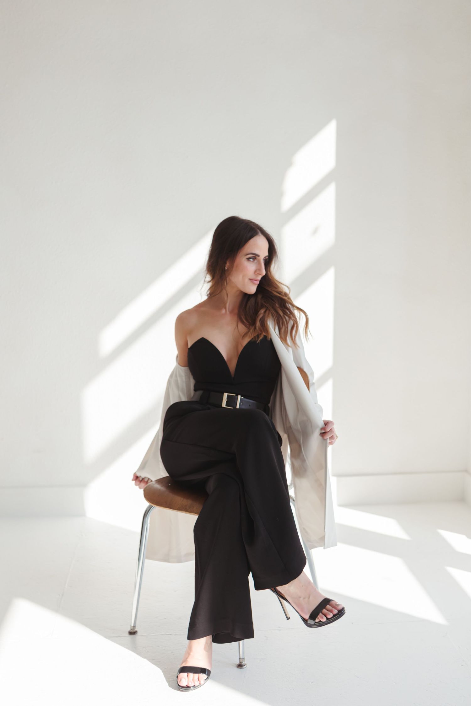A person in a black outfit poses against a white wall with dramatic shadows creating an artistic composition.