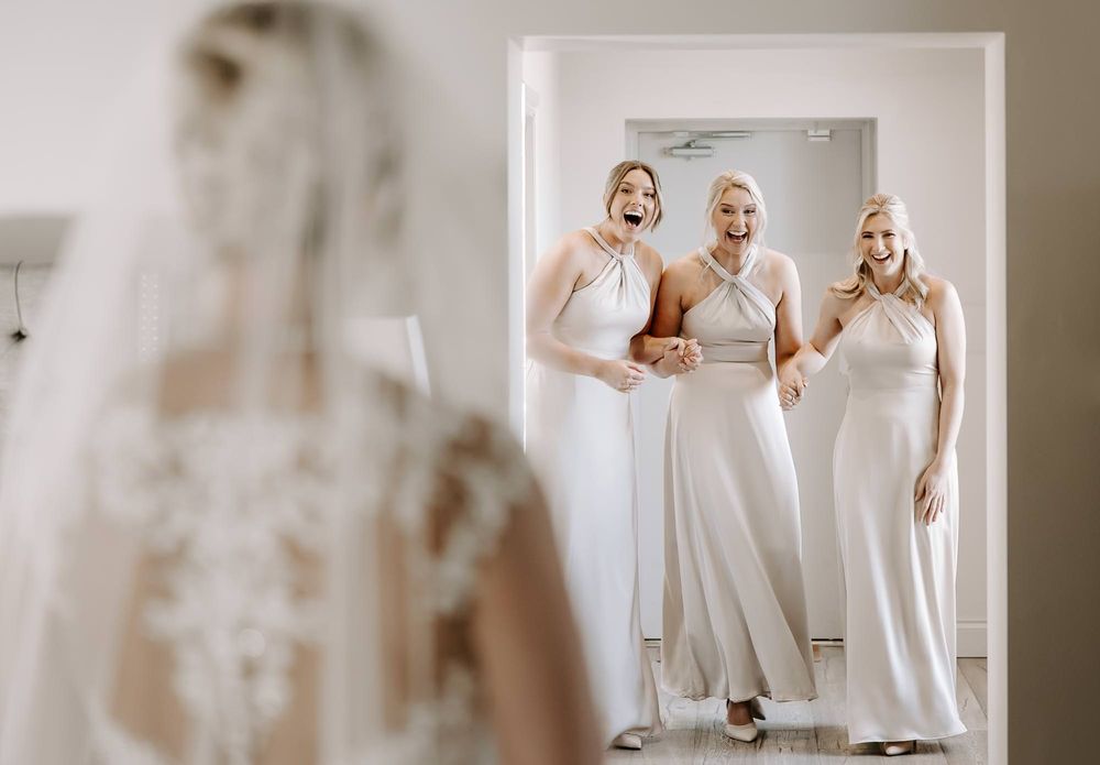 Three bridesmaids in elegant white dresses share a joyful moment while looking in a mirror at a wedding preparation.