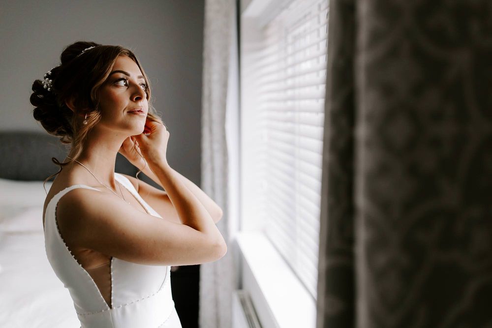 A bride in an elegant white dress adjusts her earrings while standing near a window in soft natural light.