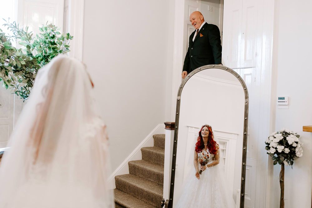 A bride in a white veil looks at her reflection in an arched mirror by a staircase with floral decorations.