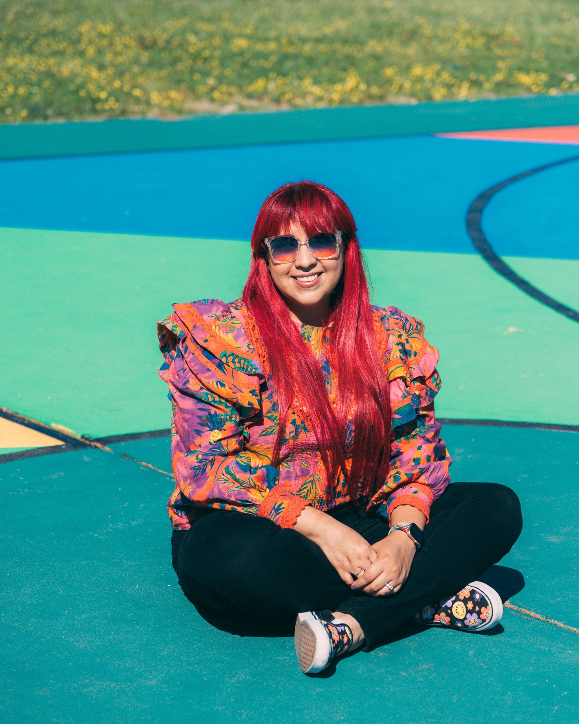 Person with vibrant red hair wearing sunglasses and a colorful floral jacket sits cross-legged on a teal outdoor surface.
