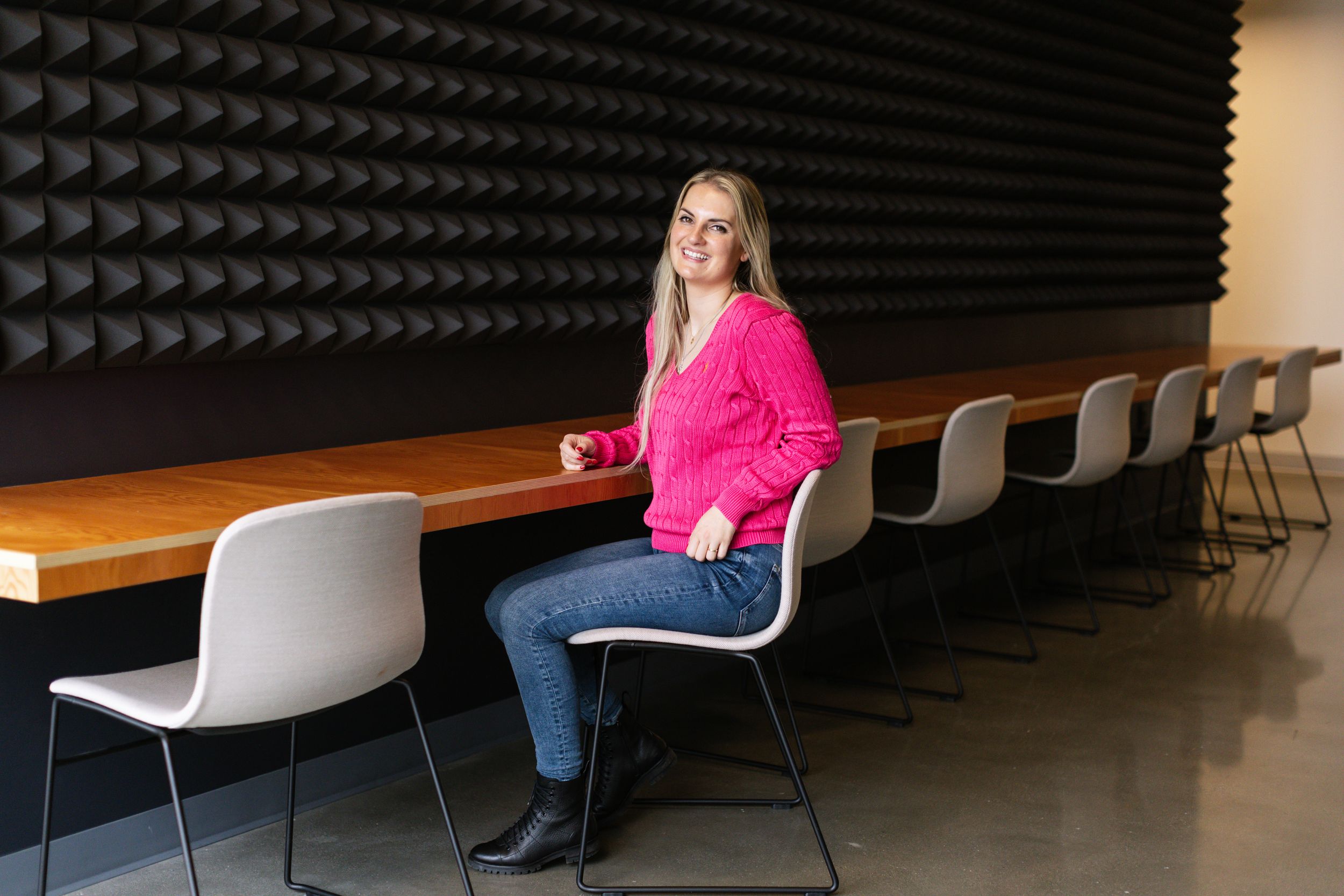 A person in a bright pink jacket sits at a wooden table in a dark room with acoustic foam panels on the wall.