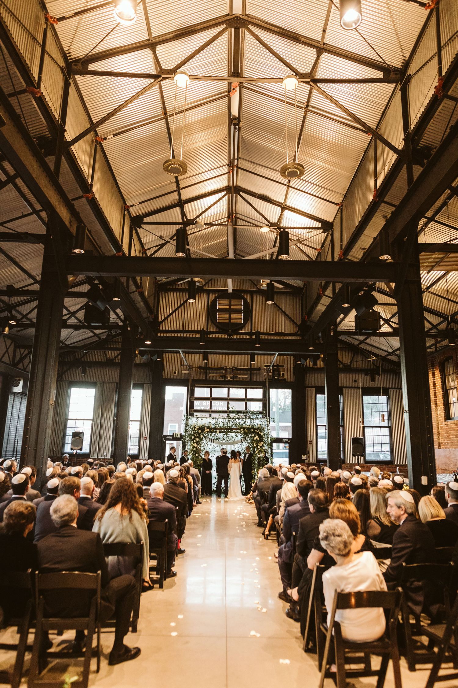 A wedding ceremony takes place in a spacious industrial venue with exposed wooden beams and warm lighting. Kehoe Iron Works, Savannah, GA