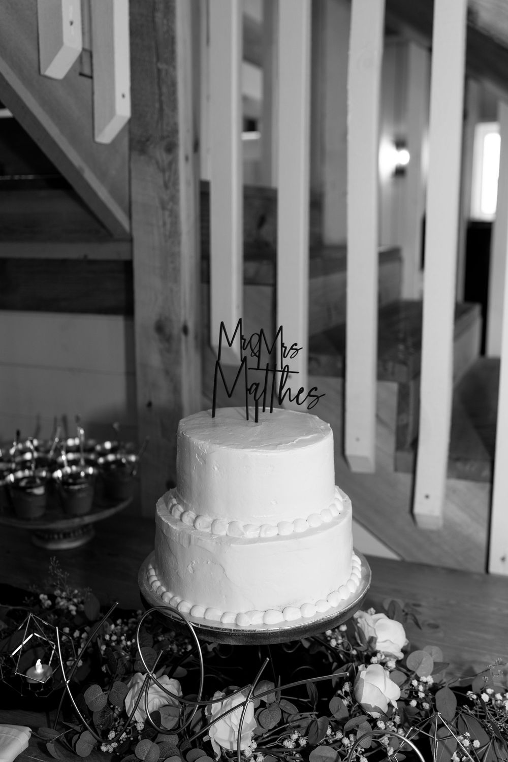 Black and white photo of two-tier white wedding cake with elegant script cake topper.