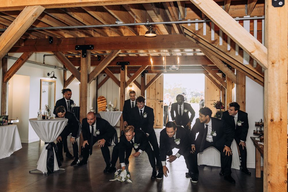Groomsmen in black suits striking fun poses together inside a rustic barn wedding venue.