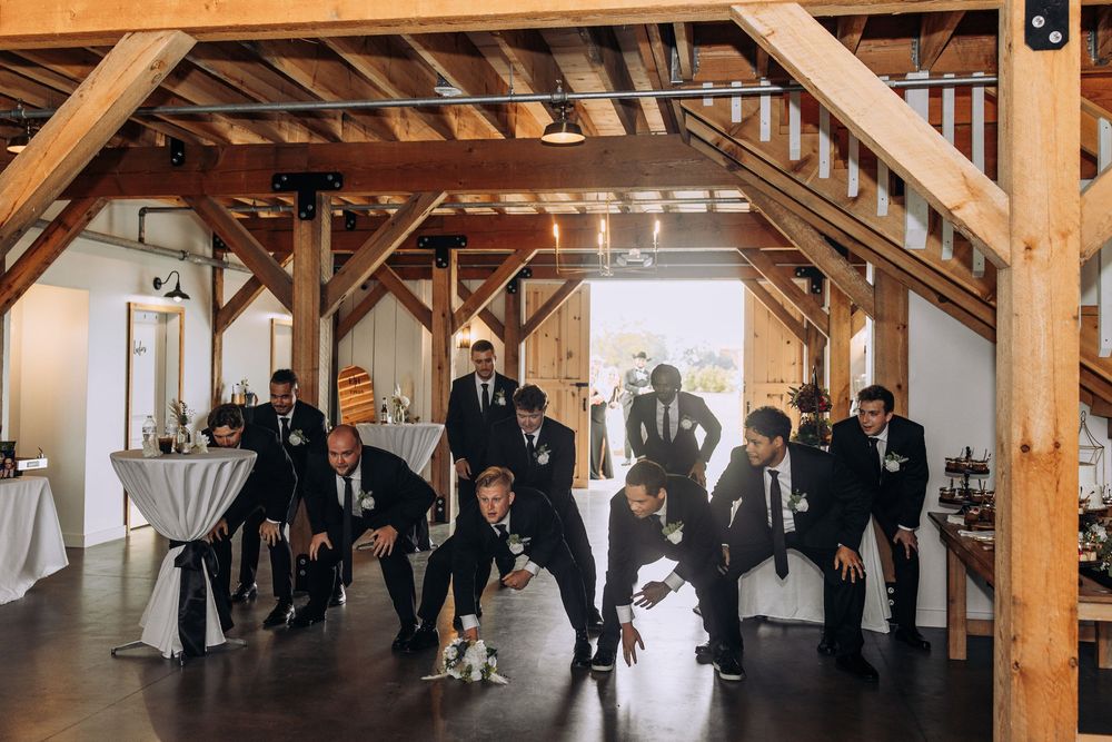 Wedding party group posing with bride in rustic barn venue with exposed wooden beam ceiling.