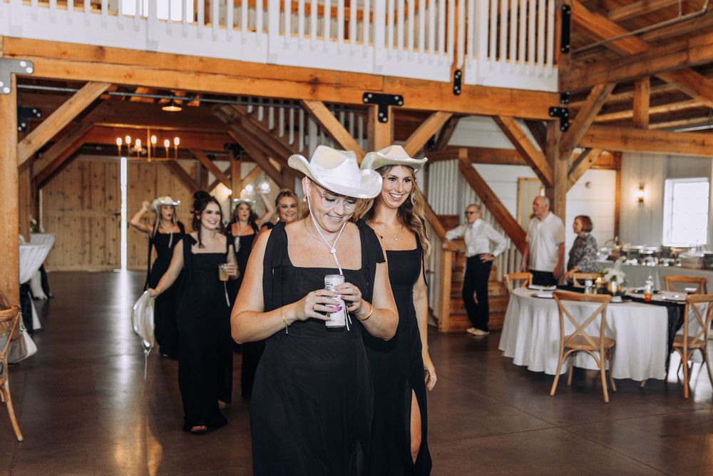 Bridesmaids in black dresses and white cowboy hats walk through a rustic barn wedding venue.