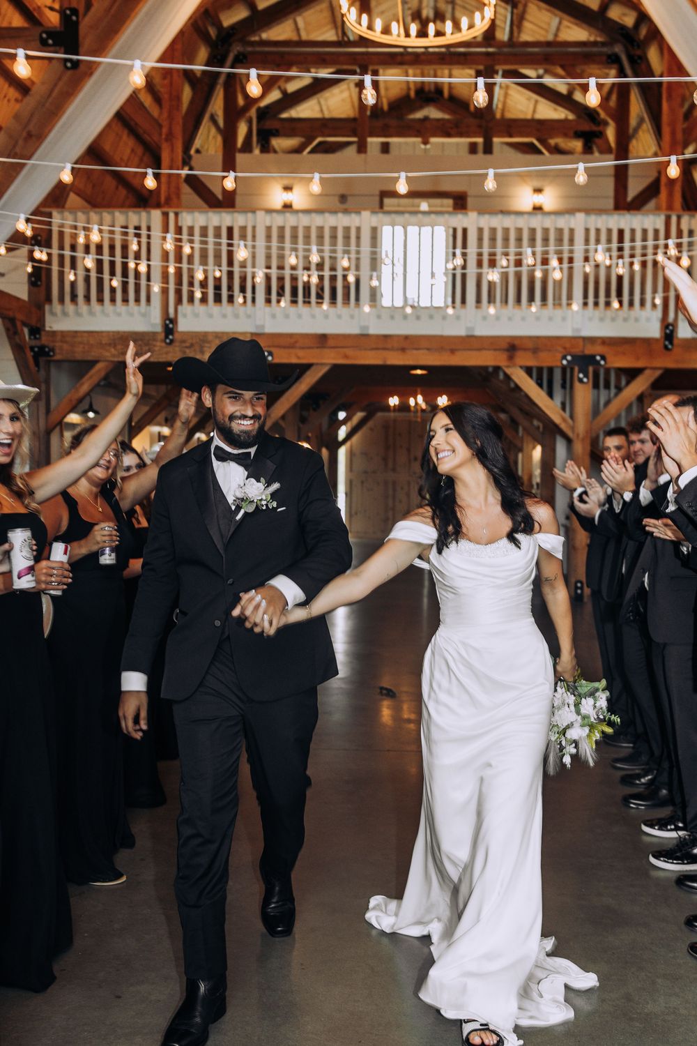 Smiling bride and groom walk hand-in-hand during grand entrance at elegant barn wedding.