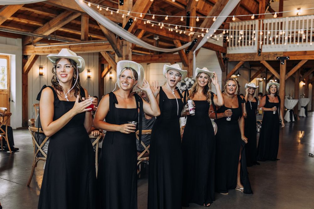 Bridesmaids in matching black gowns and white cowboy hats celebrate at a rustic barn wedding.