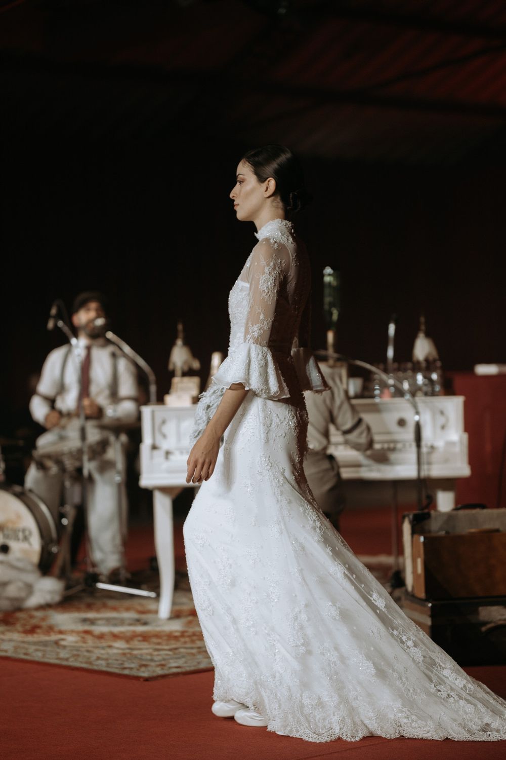 A bride in a flowing white lace wedding gown stands in a rustic indoor venue with a red wall in the background.