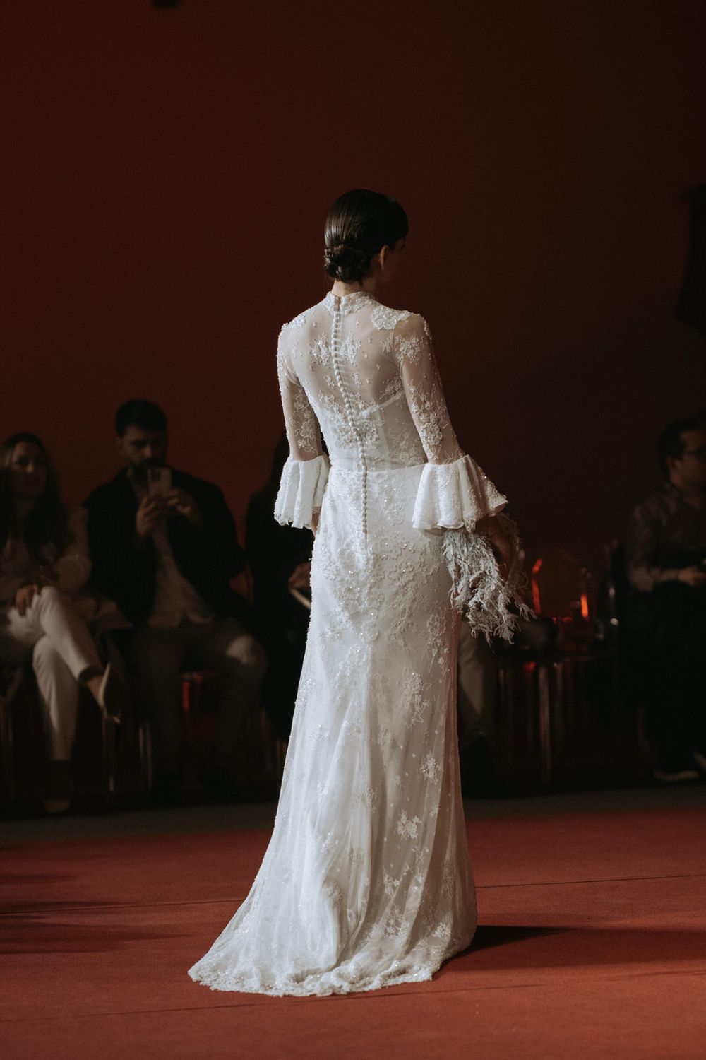 Elegant lace wedding gown with bell sleeves and button-up back shown from behind on red runway during fashion show.