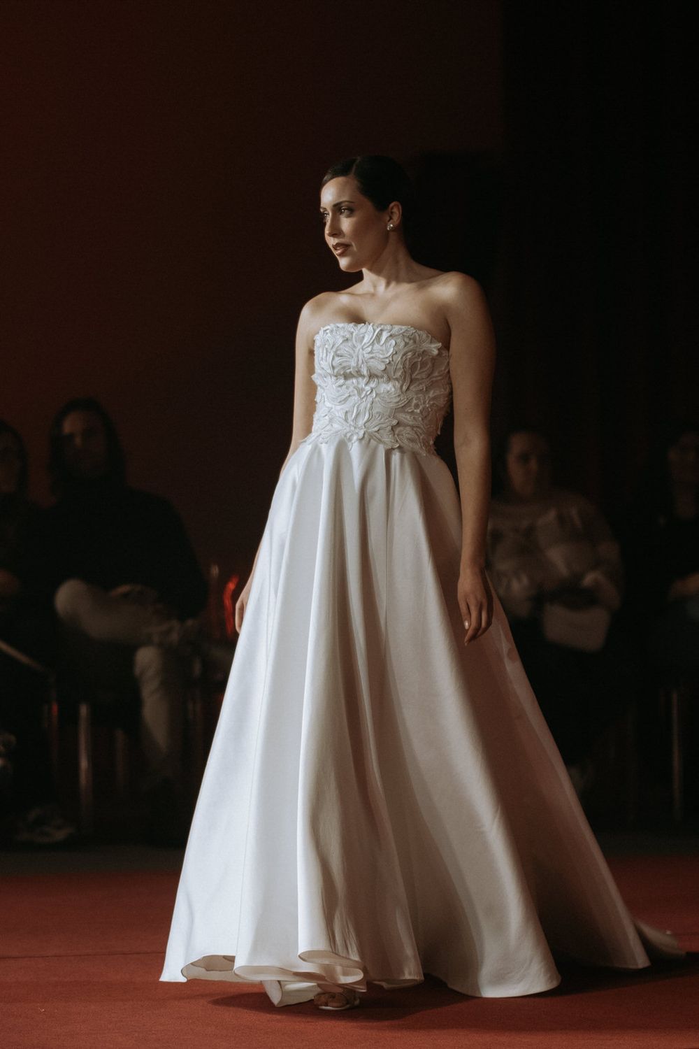 A model walks the runway in a white strapless empire waist wedding gown with delicate lace details.