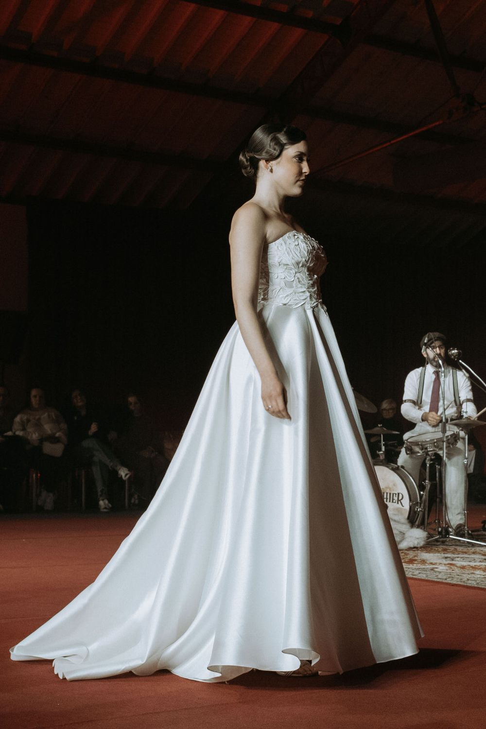 Models showcase elegant white wedding gowns with flowing trains on a rustic red carpet during a bridal fashion show.