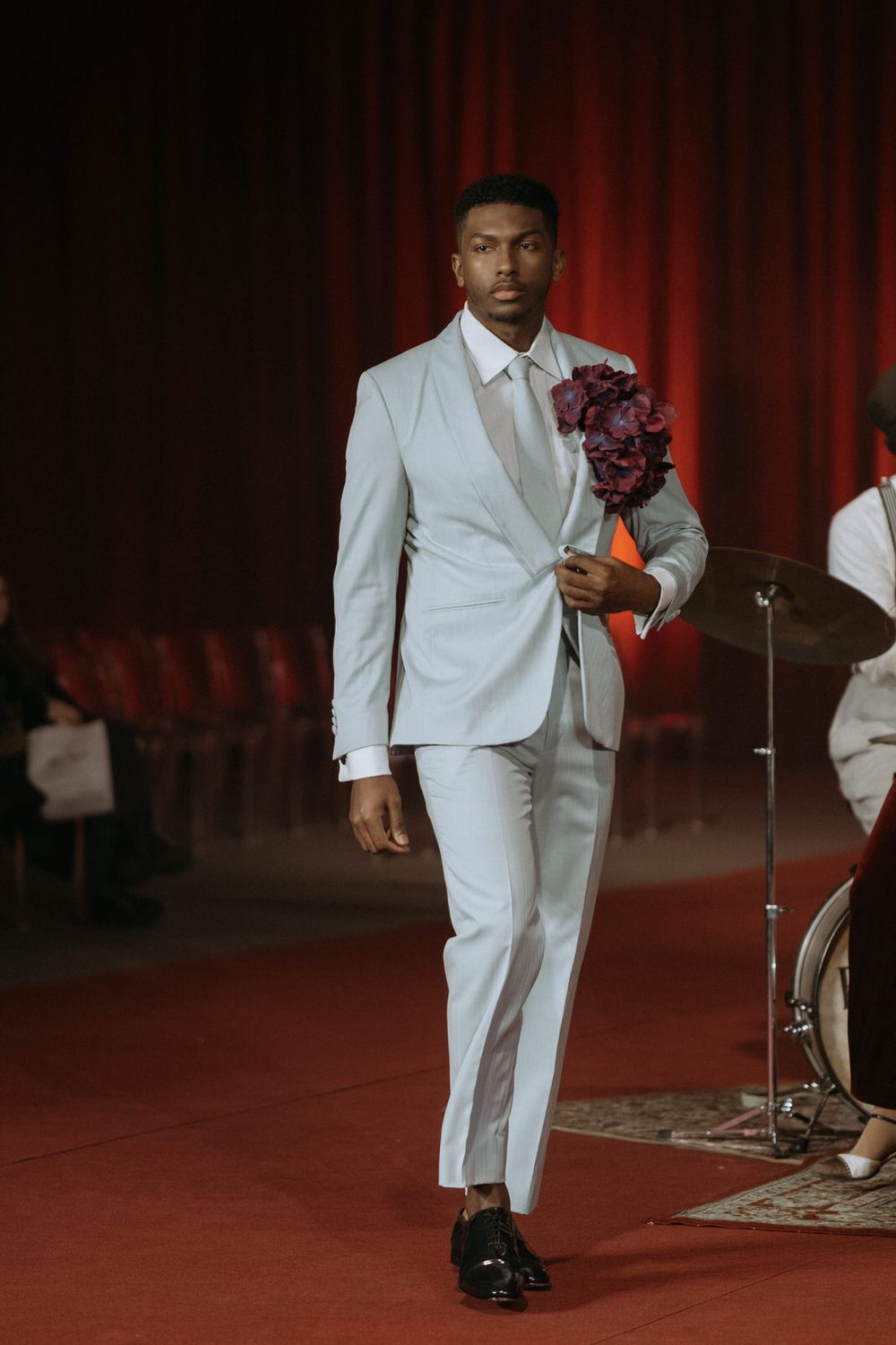 A performer in a crisp white suit walks across a stage with red curtains and musical instruments in the background.