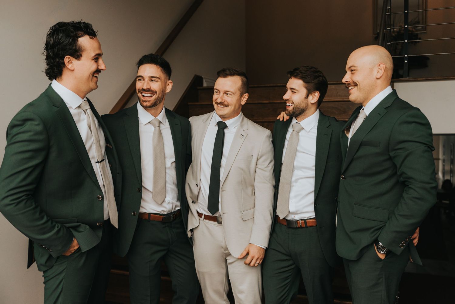 Groomsmen in green suits and khaki pants share candid laughing moments during wedding party photos.