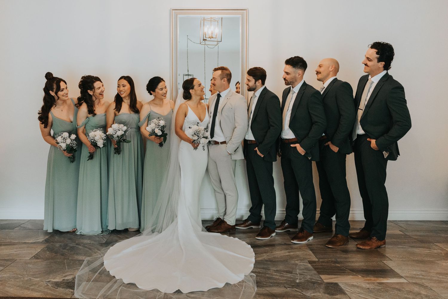 Wedding party members in sage green dresses and dark suits stand in a line against a white wall for formal portraits.
