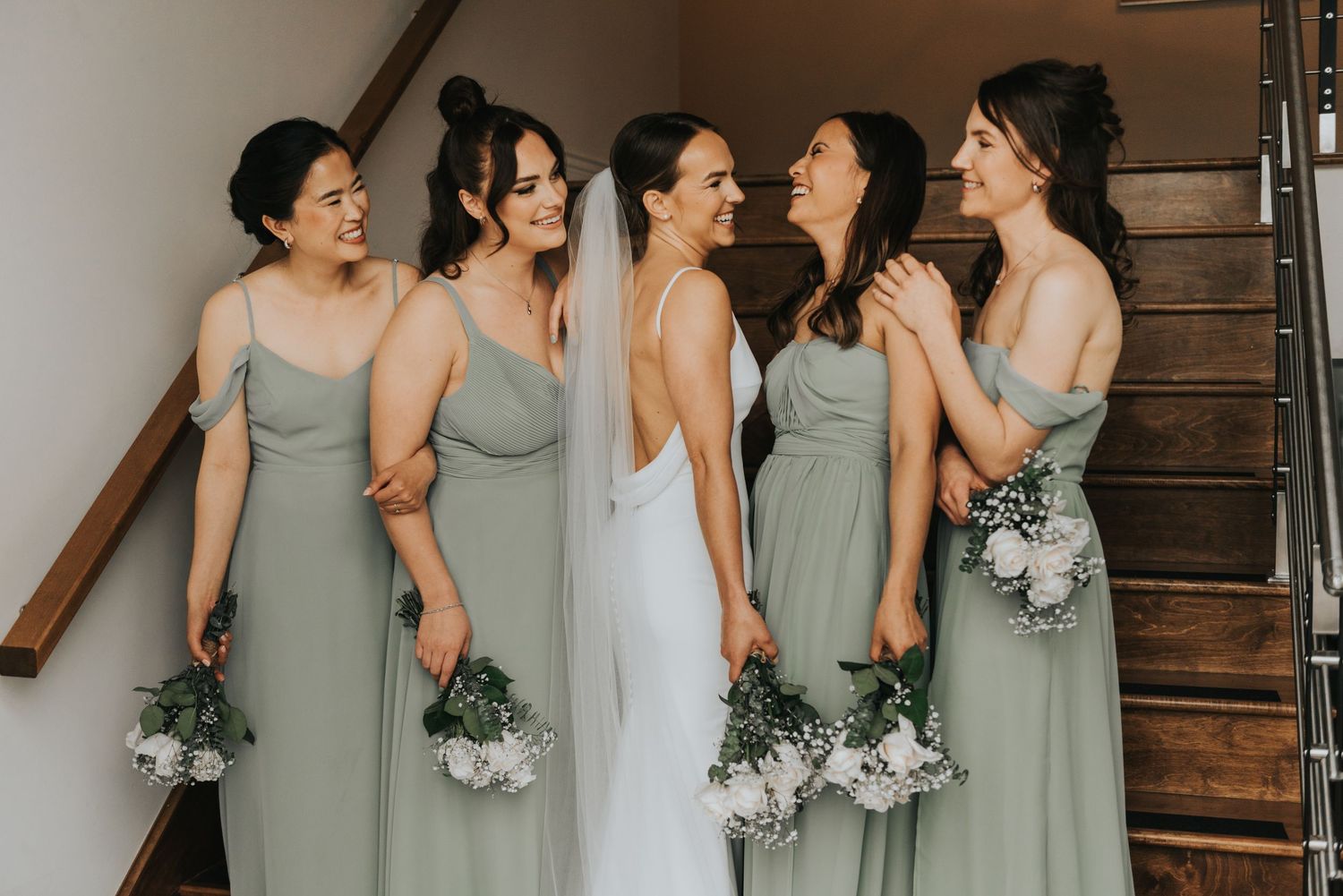 Bridesmaids in sage green dresses and white bouquets share candid moments along rustic wooden staircase.