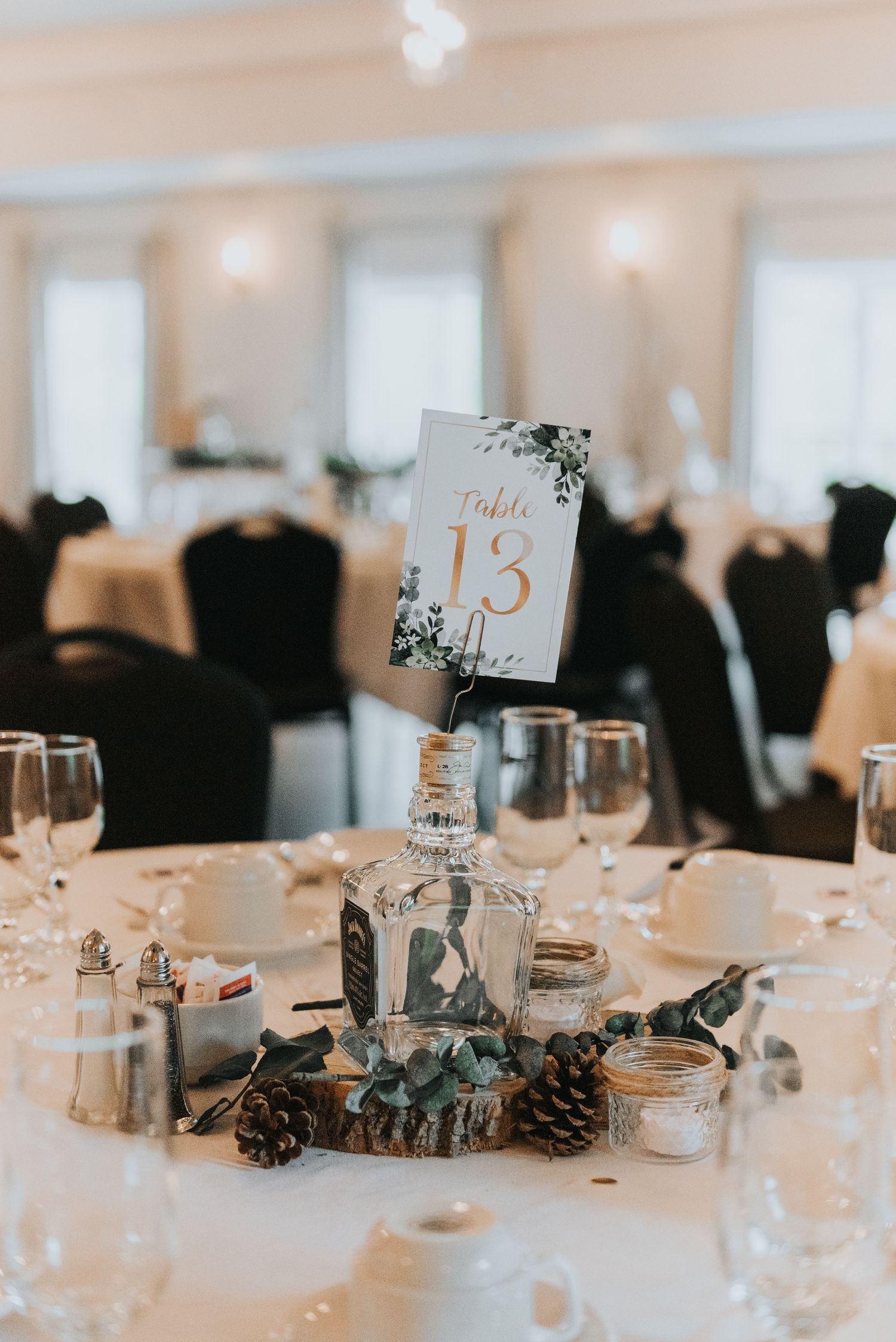 Wedding reception table setting with table number 15, pinecones, and wine glasses on white tablecloth in elegant venue.