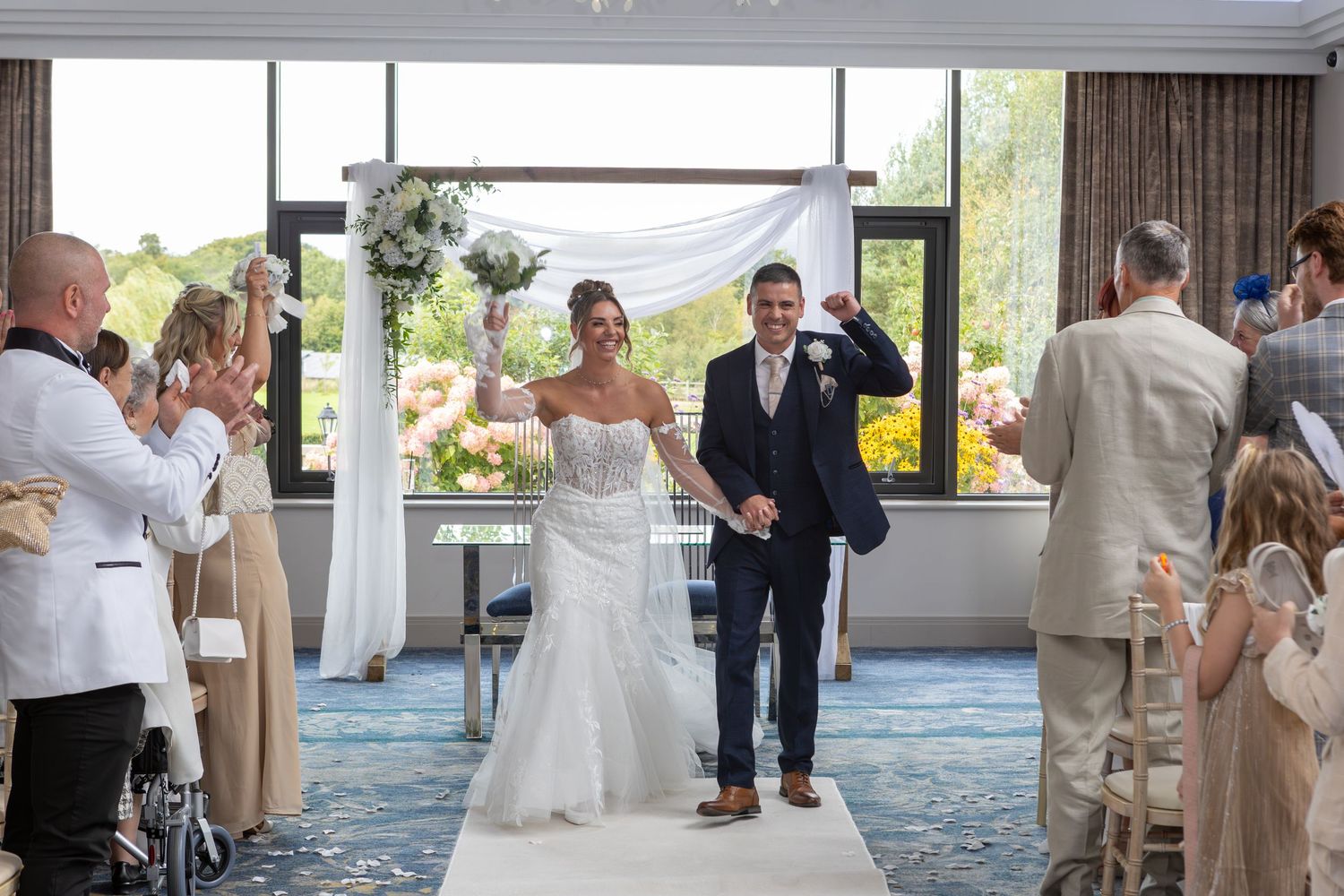 A happy couple makes their grand entrance at a wedding reception while guests applaud in a bright modern venue.