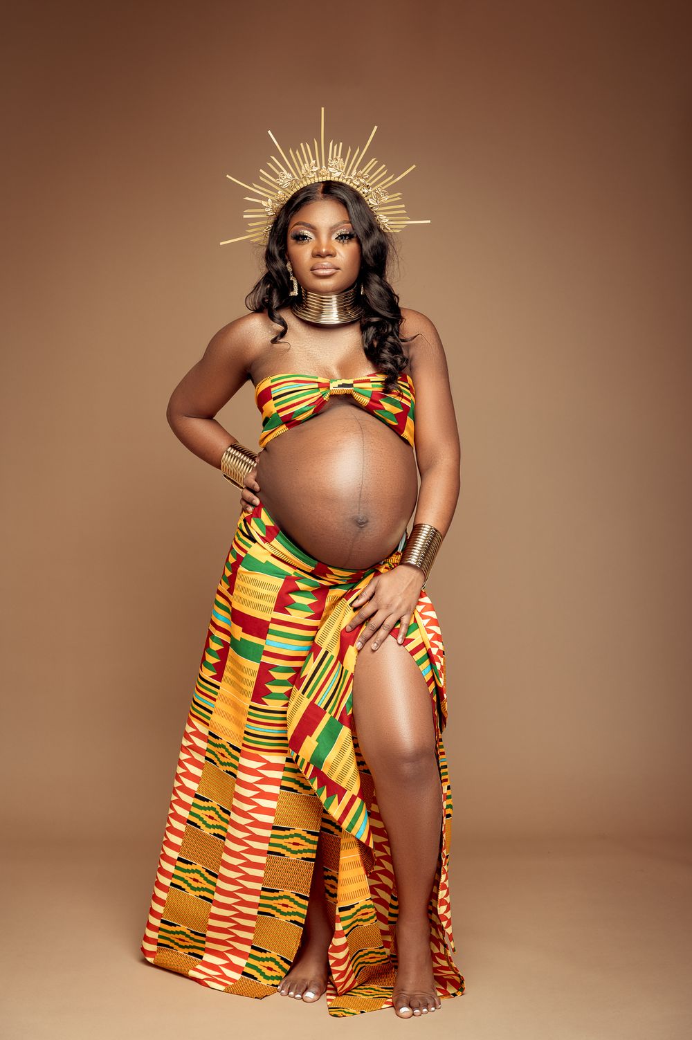 A maternity photoshoot featuring vibrant kente print fabric and gold accessories against a warm brown backdrop.