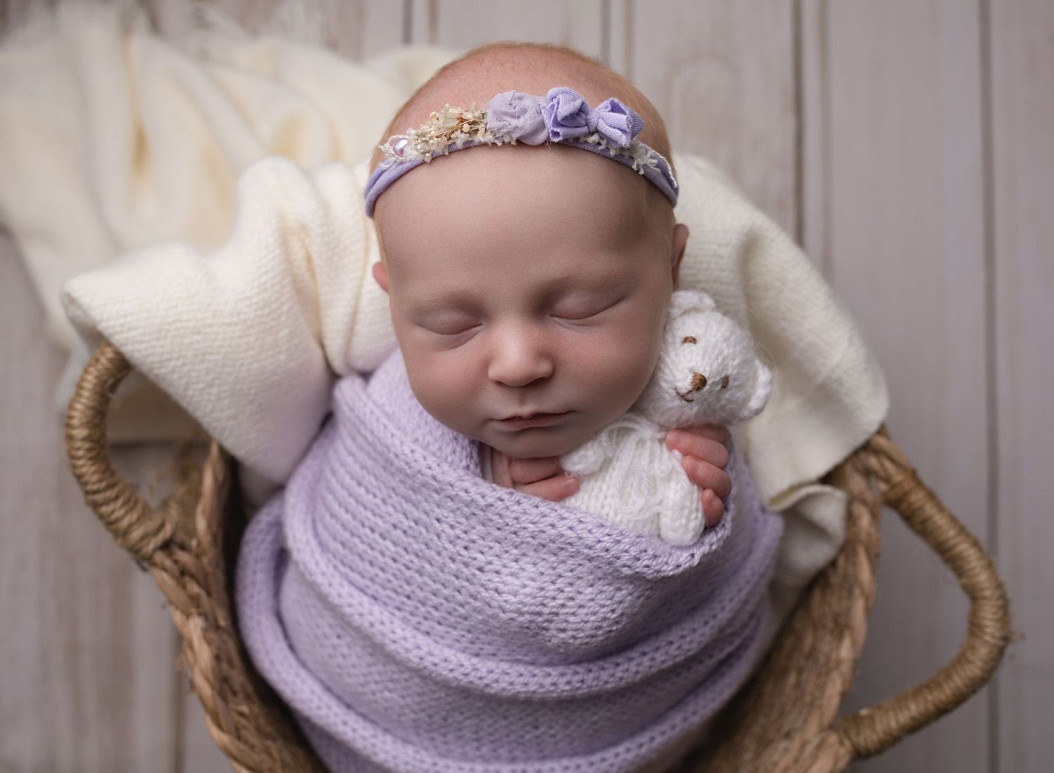 sleeping baby girl wrapped in lilac and posed gently in a wicker basket holds small white teddy, newborn photography session