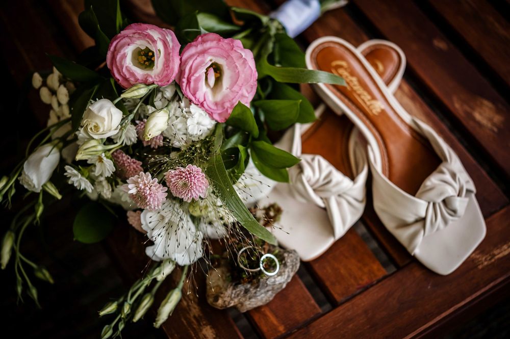 Wedding details including pink roses, white shoes, and gold rings arranged on wooden surface.
