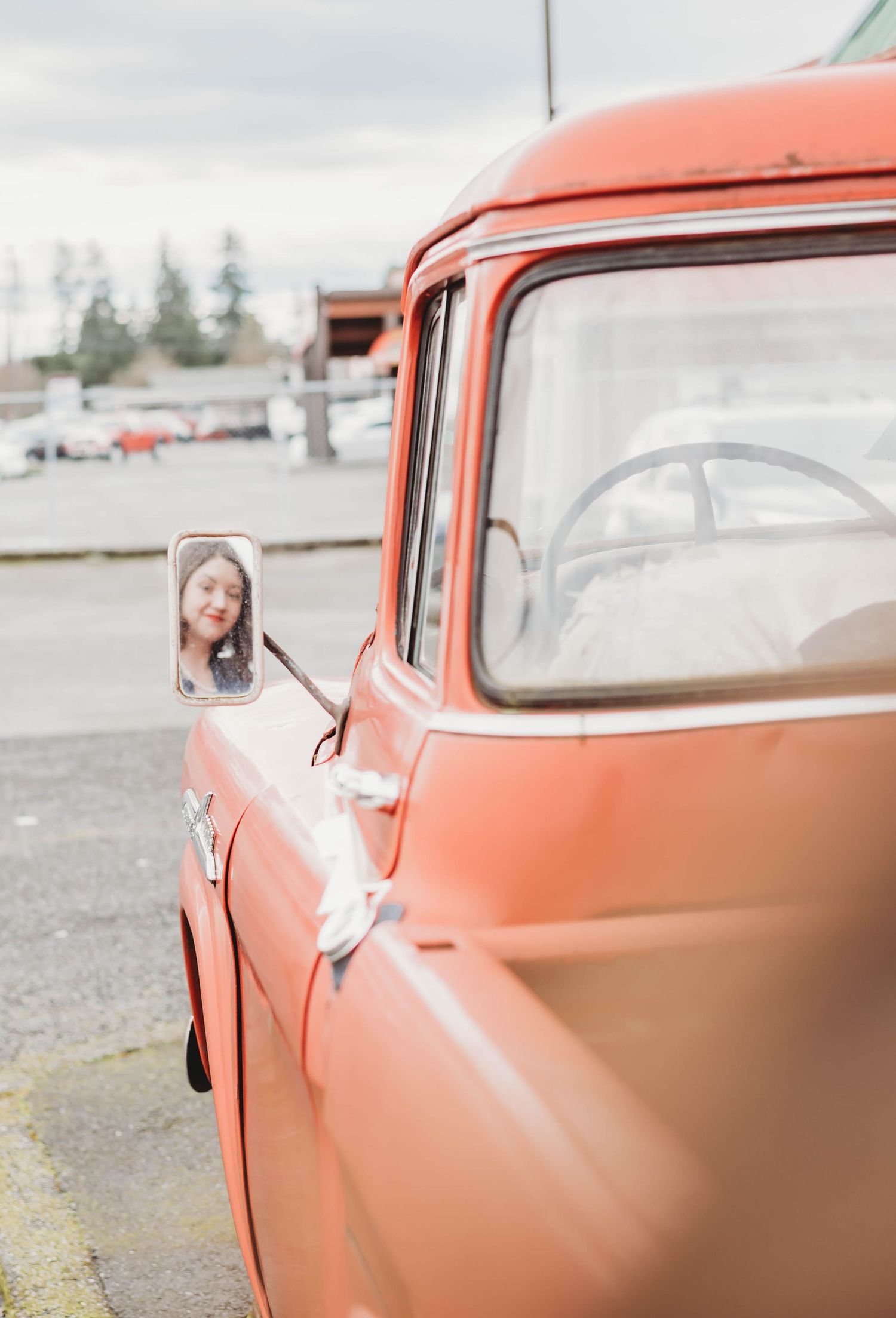 A vintage coral-colored truck with side mirror detail parked in a gravel lot on an overcast day.