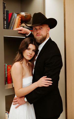 Couple in wedding attire share a tender moment by a bookshelf, with him in a western-style hat.