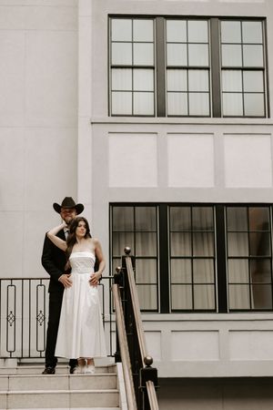 Wedding couple embraces on outdoor steps with black railings and white walls.
