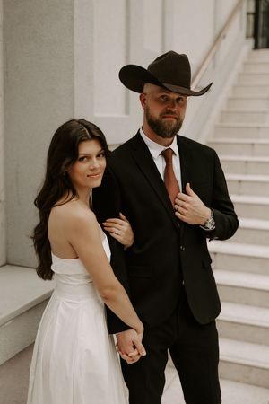 A western-themed wedding portrait featuring formal attire and cowboy hat against white architecture.