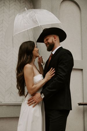 A couple embraces tenderly under a white umbrella in front of a textured wall.