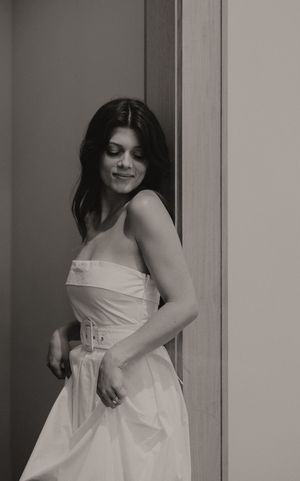 A person in a white dress poses against a light colored wall.
