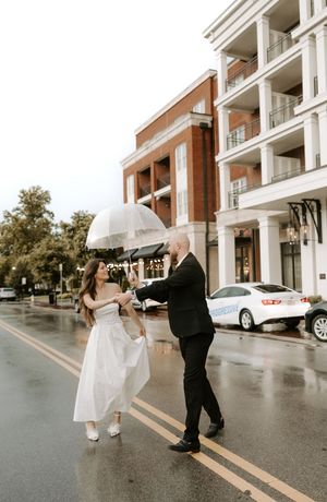 A couple walks together on a rainy street sharing a clear umbrella outside a brick building.