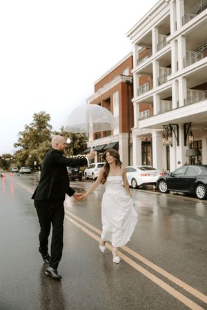 A couple dances playfully in the rain while sharing an umbrella on a wet street.