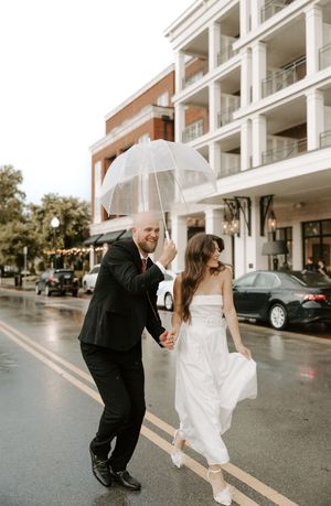 Newlyweds dance and laugh together on a rainy street with white building in background.