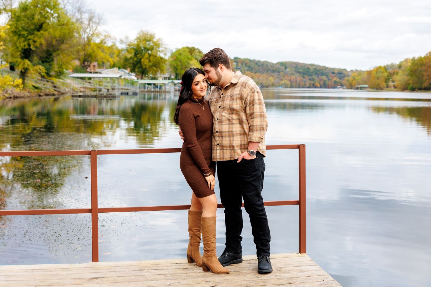 A couple stands together on a wooden dock overlooking a serene lake with autumn trees and boat docks in the background.