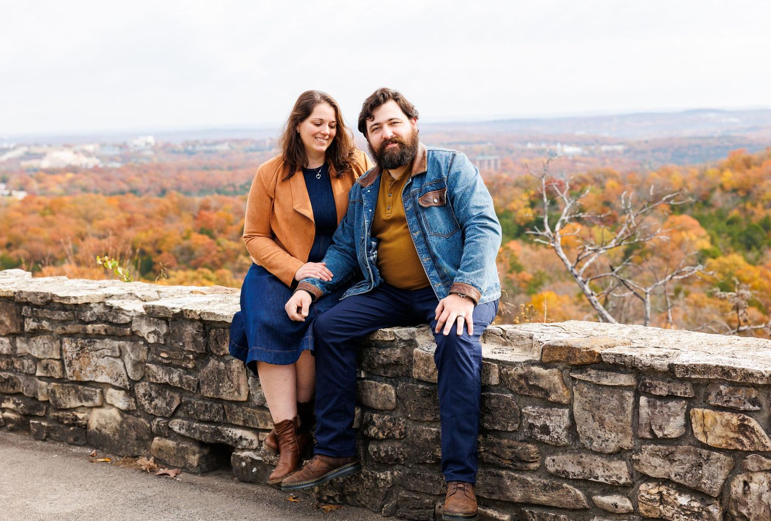 A couple stands together on a wooden dock overlooking a serene lake with autumn trees and boat docks in the background.
